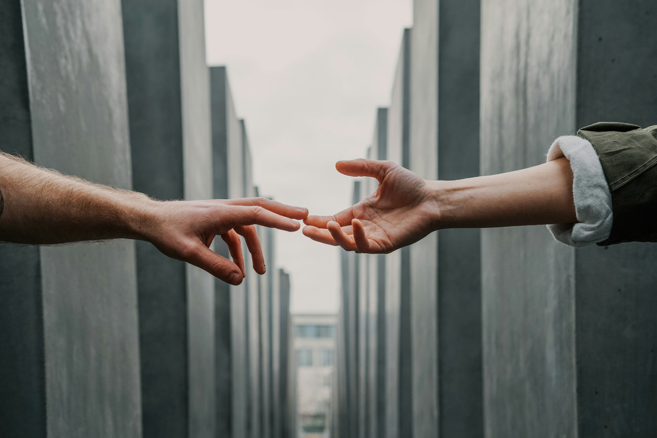 Two hands reaching out to each other between large gray stone structures in an outdoor setting.